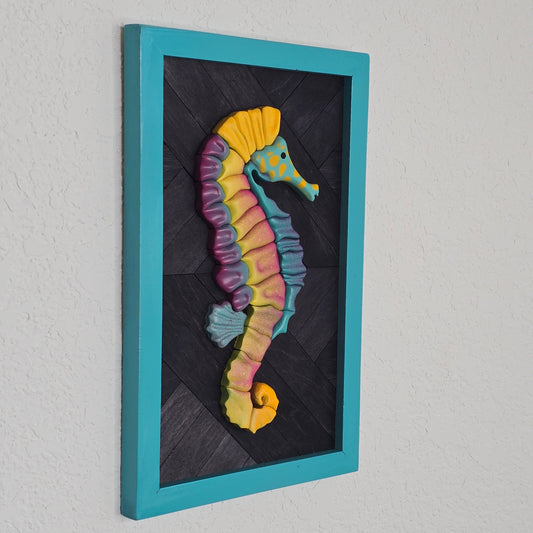 Handcrafted seahorse sculpture with neon-style color segments in purple, yellow, and blue. The piece is mounted on a dark grey, textured wooden parquet backing and enclosed in a bright turquoise rectangular frame.