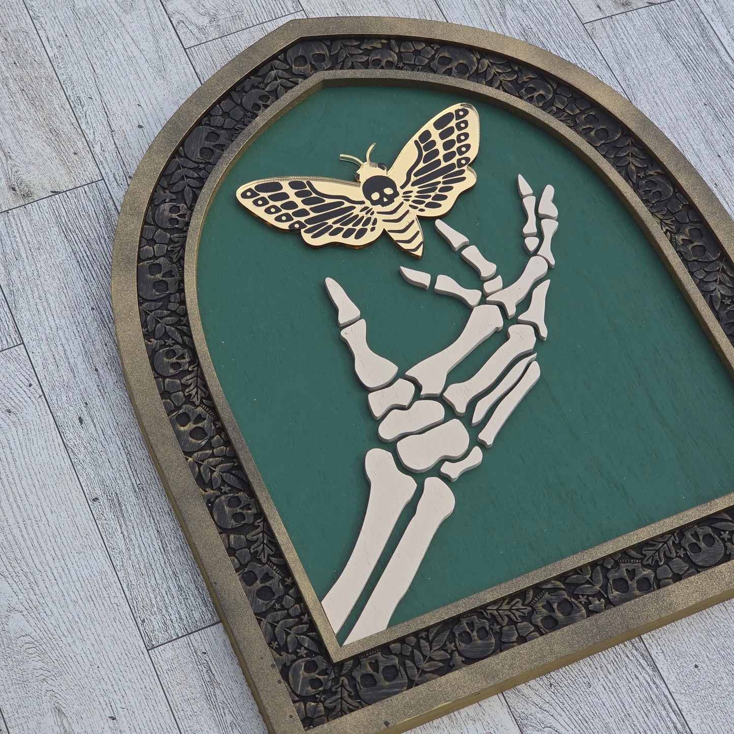 Decorative wall art piece with skeleton hand, death moth, and decorative border on a wooden background