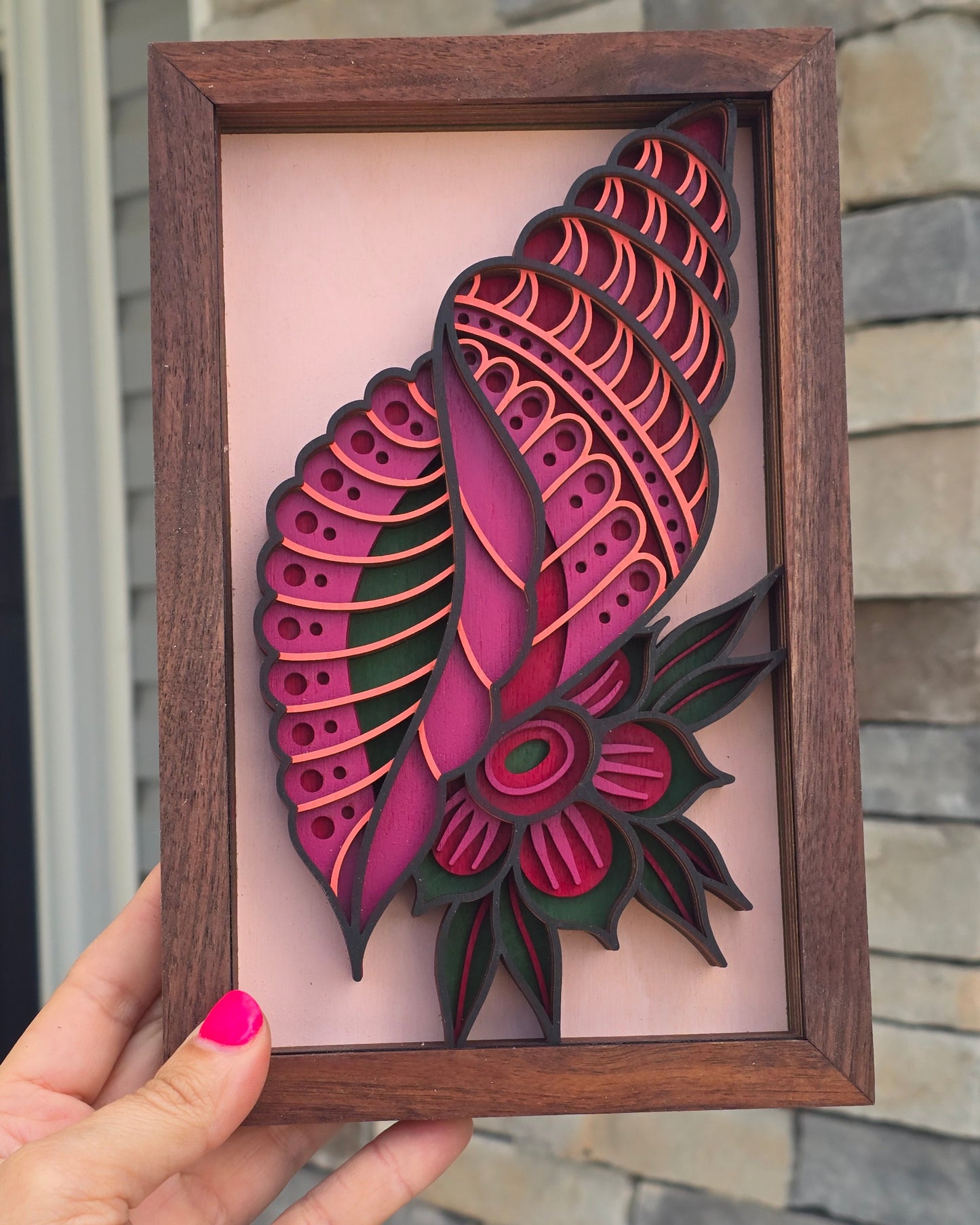 A handcrafted rectangular wood wall art piece held by a hand. The design features a 3D, multi-layered laser-cut seashell in vibrant shades of magenta, coral, and deep hunter green. A matching geometric flower blooms from the side of the shell against a soft pink background, enclosed in a dark wood frame.
