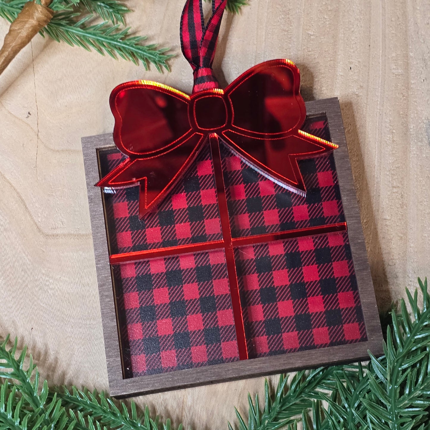 Red and black plaid gift box with a red bow on a wooden surface with greenery.