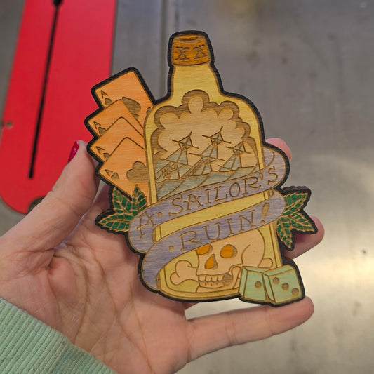 Hand-painted wooden magnet art of "A Sailor's Ruin" featuring a ship in a bottle, playing cards, dice, and a skull in a vintage tattoo aesthetic.