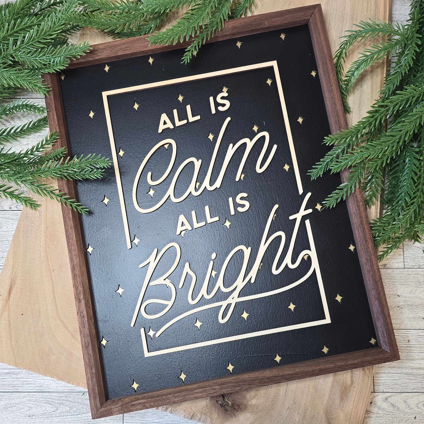 Decorative sign with 'All is Calm, All is Bright' text on a wooden surface with greenery.