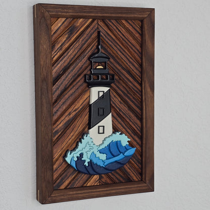 Handcrafted 3D wooden lighthouse art featuring a black and white striped tower and a detailed blue and light teal cresting wave. The piece is mounted horizontally on a rich, medium-brown wood inlay with a diagonal chevron pattern, set in a matching dark wood frame.