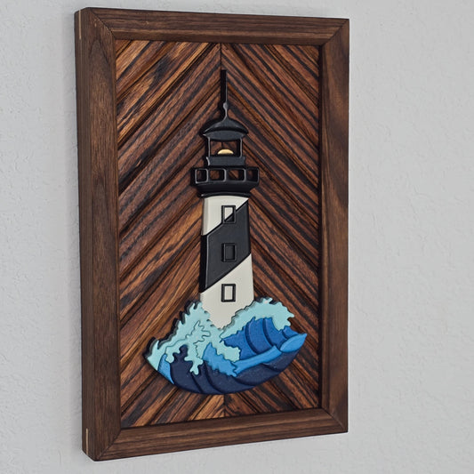 Handcrafted 3D wooden lighthouse art featuring a black and white striped tower and a detailed blue and light teal cresting wave. The piece is mounted horizontally on a rich, medium-brown wood inlay with a diagonal chevron pattern, set in a matching dark wood frame.