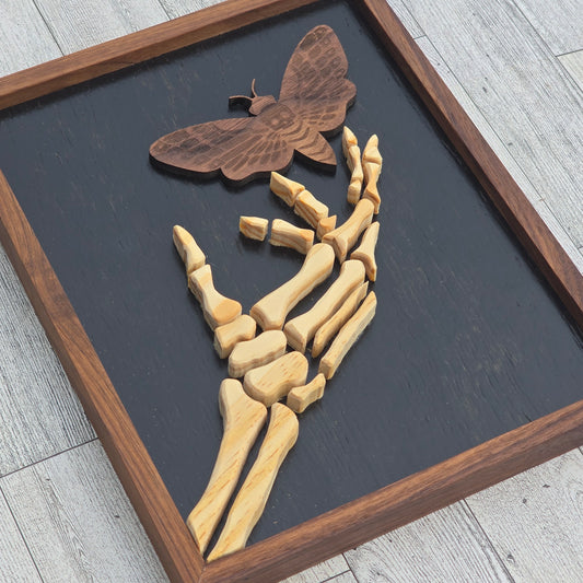 Wooden skeleton hand holding a butterfly on a dark background with a wooden frame.