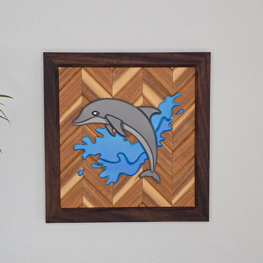A handcrafted wooden wall piece featuring a grey dolphin leaping out of bright blue splashing water. The artwork is mounted on a light-toned wood herringbone pattern and enclosed in a square, dark walnut-colored frame.