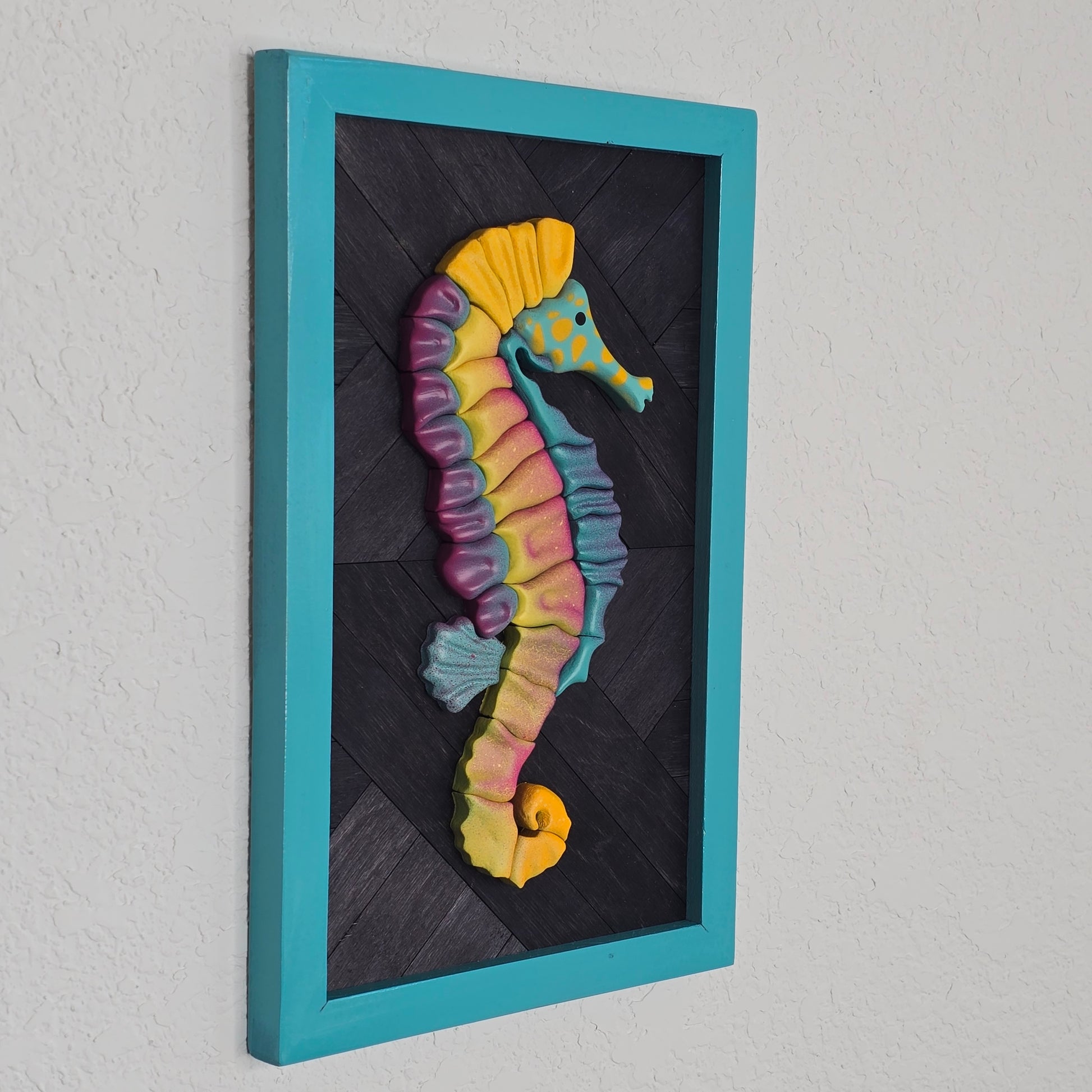 Handcrafted seahorse sculpture with neon-style color segments in purple, yellow, and blue. The piece is mounted on a dark grey, textured wooden parquet backing and enclosed in a bright turquoise rectangular frame.