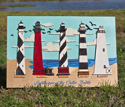 Five Lighthouses made from wood and painted to look like the five lighthouses of the outer banks, on a backer piece that resembles the beach and the sky. Placed on the ground in green grass