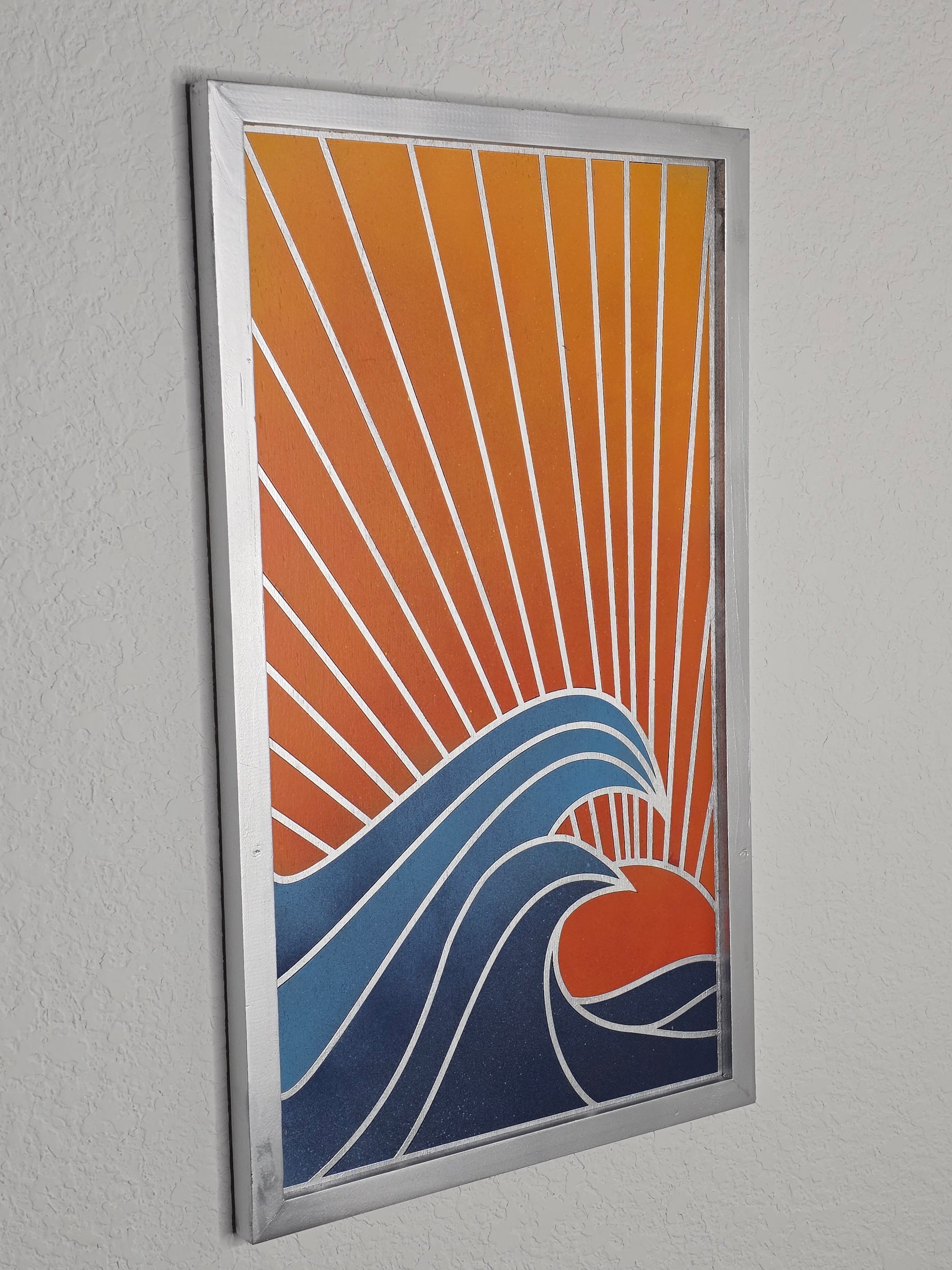 A vertical wood art piece titled Ocean Sunset. A red sun sets behind large, curling blue waves with white outlines. Diagonal rays fill the orange sky, enclosed in a silver frame.
