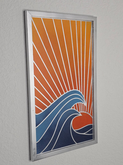 A vertical wood art piece titled Ocean Sunset. A red sun sets behind large, curling blue waves with white outlines. Diagonal rays fill the orange sky, enclosed in a silver frame.