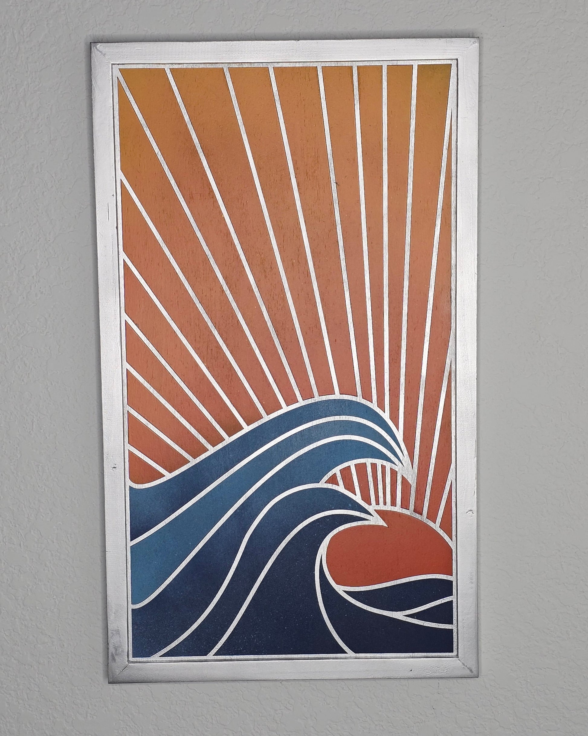 A vertical wood art piece titled Ocean Sunset. A red sun sets behind large, curling blue waves with white outlines. Diagonal rays fill the orange sky, enclosed in a silver frame.
