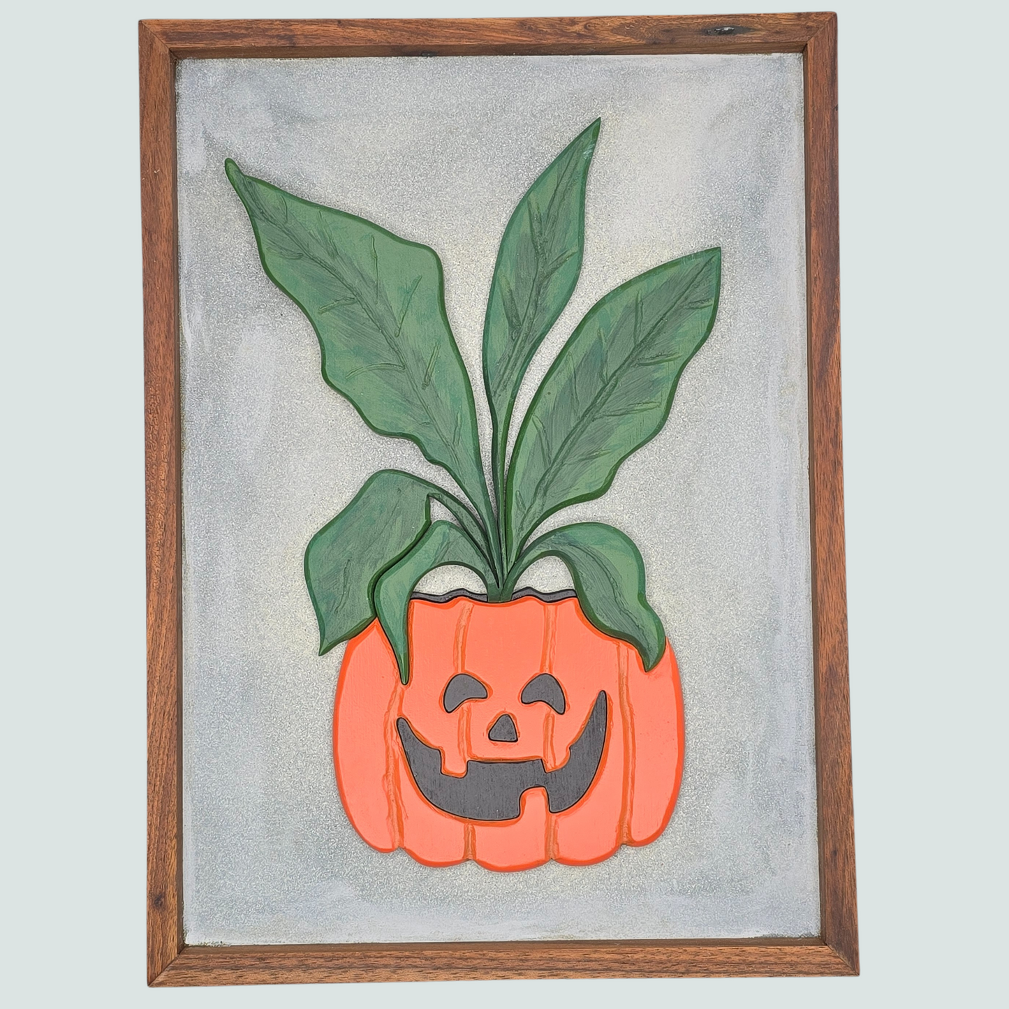 Framed artwork of a pumpkin with a face and green leaves on a light gray background