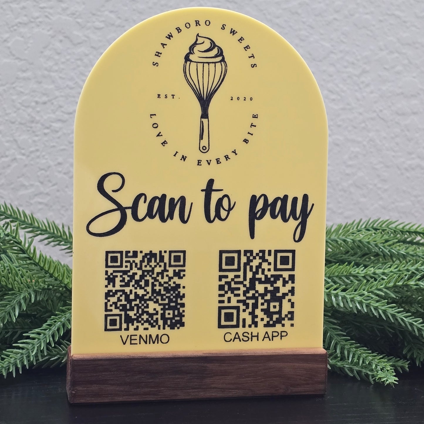 Yellow sign with QR codes for Cash App and Venmo, featuring a logo and text, against a gray wall with greenery. Branded for Shawboro Sweets