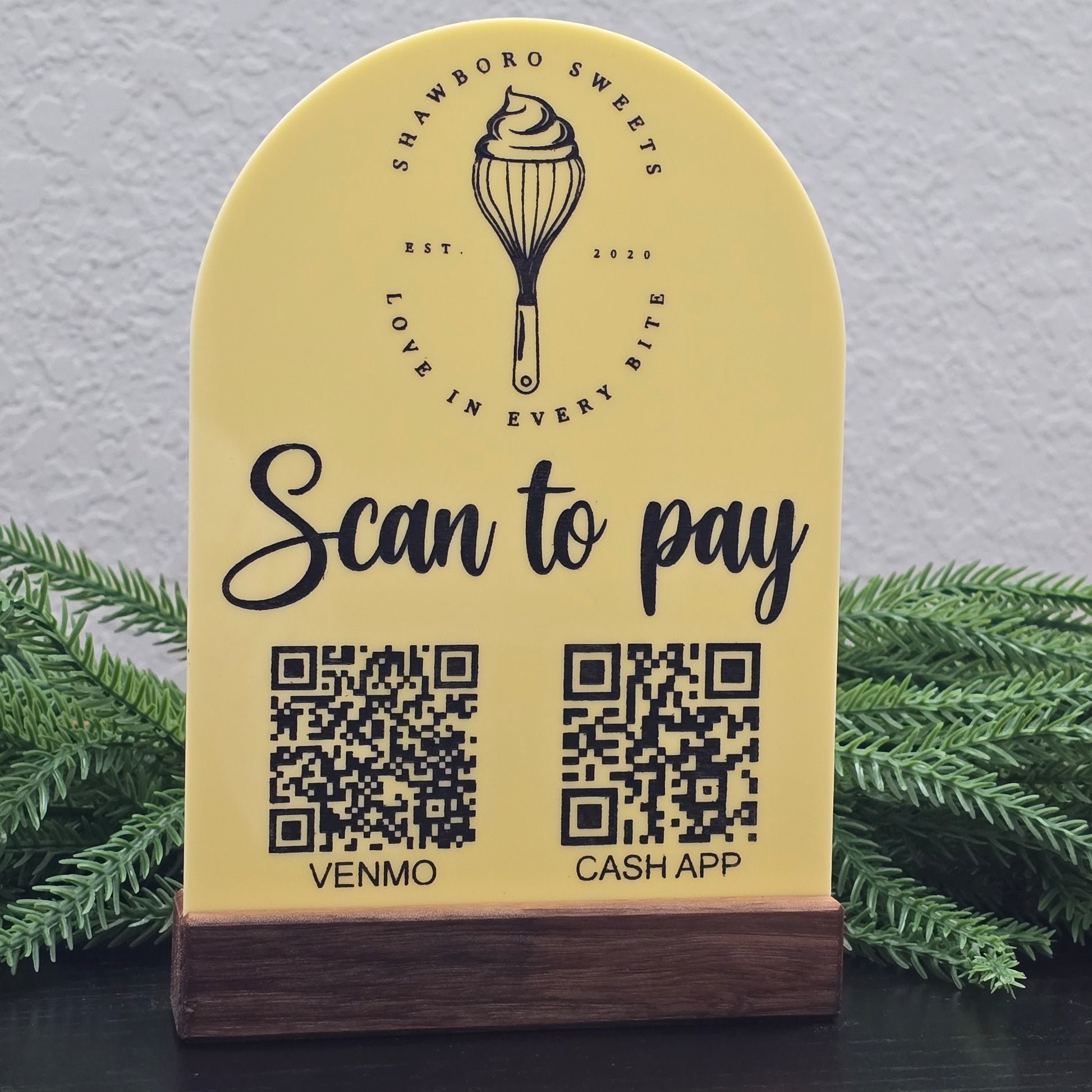Yellow sign with QR codes for Cash App and Venmo, featuring a logo and text, against a gray wall with greenery. Branded for Shawboro Sweets