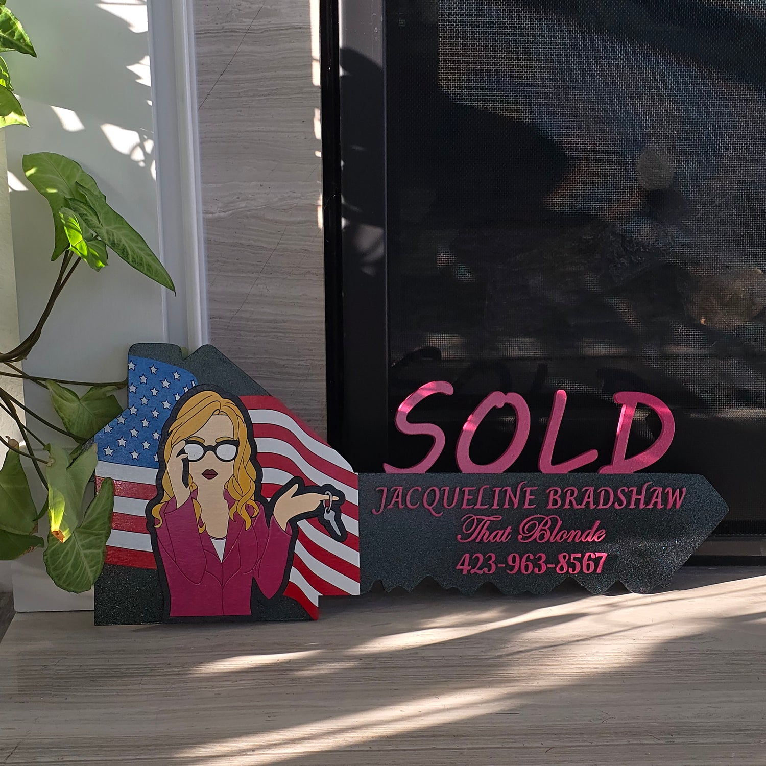 Real estate 'SOLD' sign with a person holding an American flag, brand name, and contact information.
