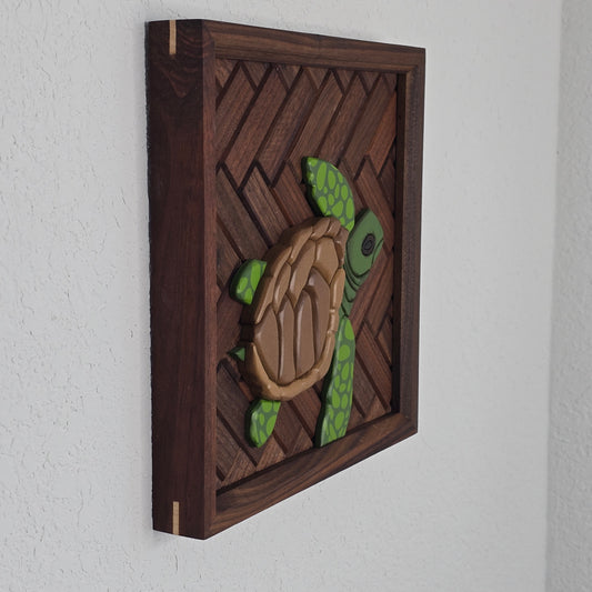 A handcrafted 3D sea turtle sculpture featuring a segmented brown shell and lime-green flippers with dark green spots. The turtle is set against a dark brown wood inlay in a diagonal basket-weave pattern, enclosed in a matching dark square frame.
