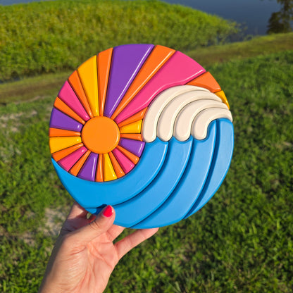 A carved art work circle where the sun rays and waves make a ying yang design. Blue waves with white caps. Sun has rays of orange, yellow, pink, and purple. 