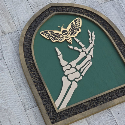 Decorative wall art piece with skeleton hand, death moth, and decorative border on a wooden background