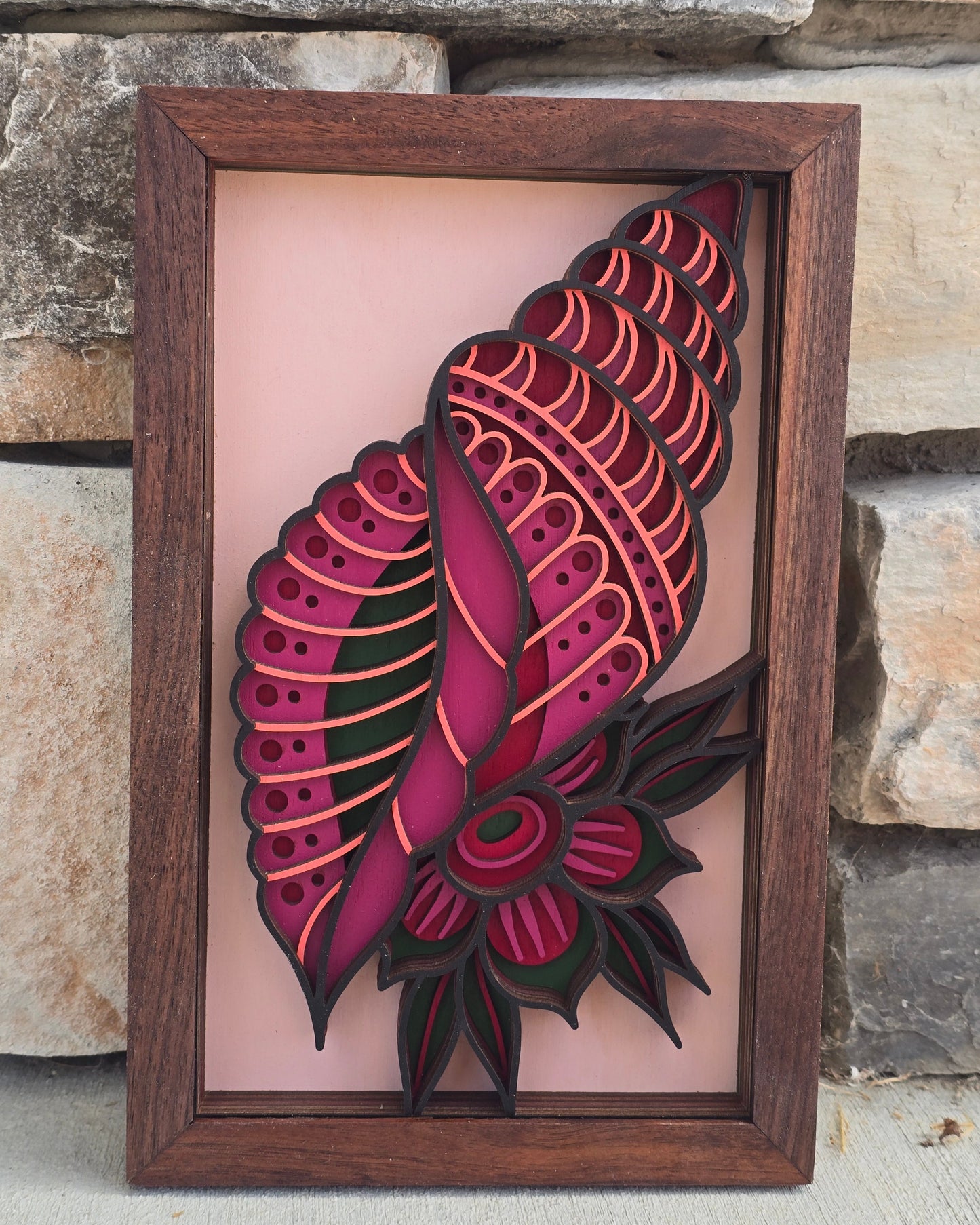 multi-layered wood art of a magenta and coral patterned seashell with a tropical flower detail inside a dark wooden frame.