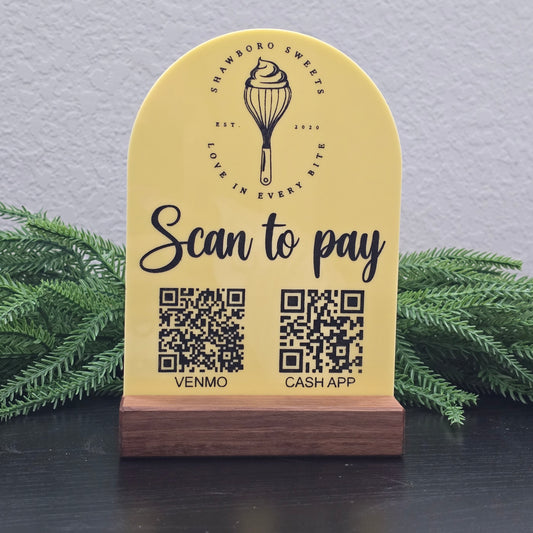 Yellow sign with QR codes for Cash App and Venmo, featuring a logo and text, against a gray wall with greenery.