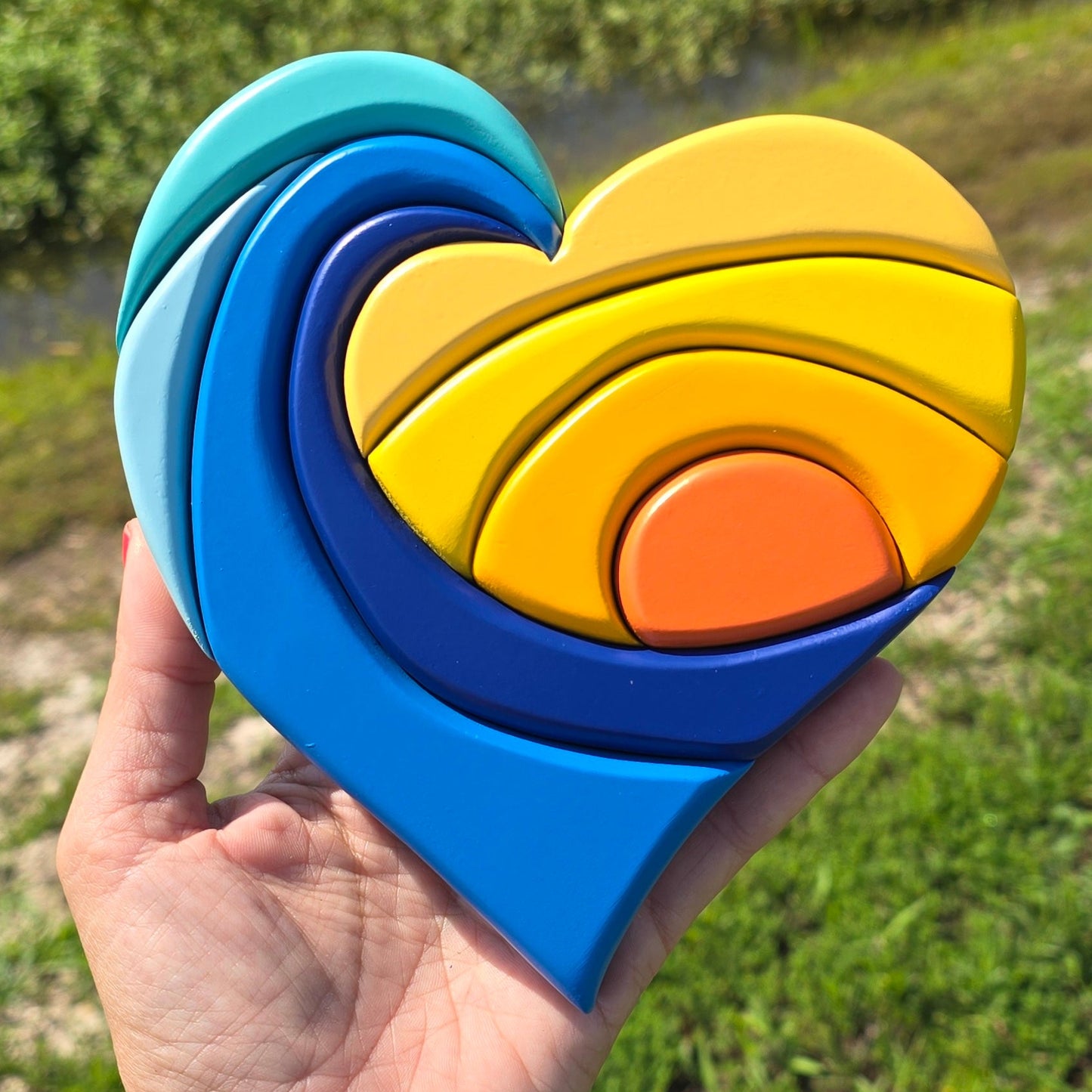 Carved wave and sunset in the shape of a heart, blues and yellows, being held with a green grass background