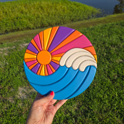 A carved art work circle where the sun rays and waves make a ying yang design. Blue waves with white caps. Sun has rays of orange, yellow, pink, and purple. 