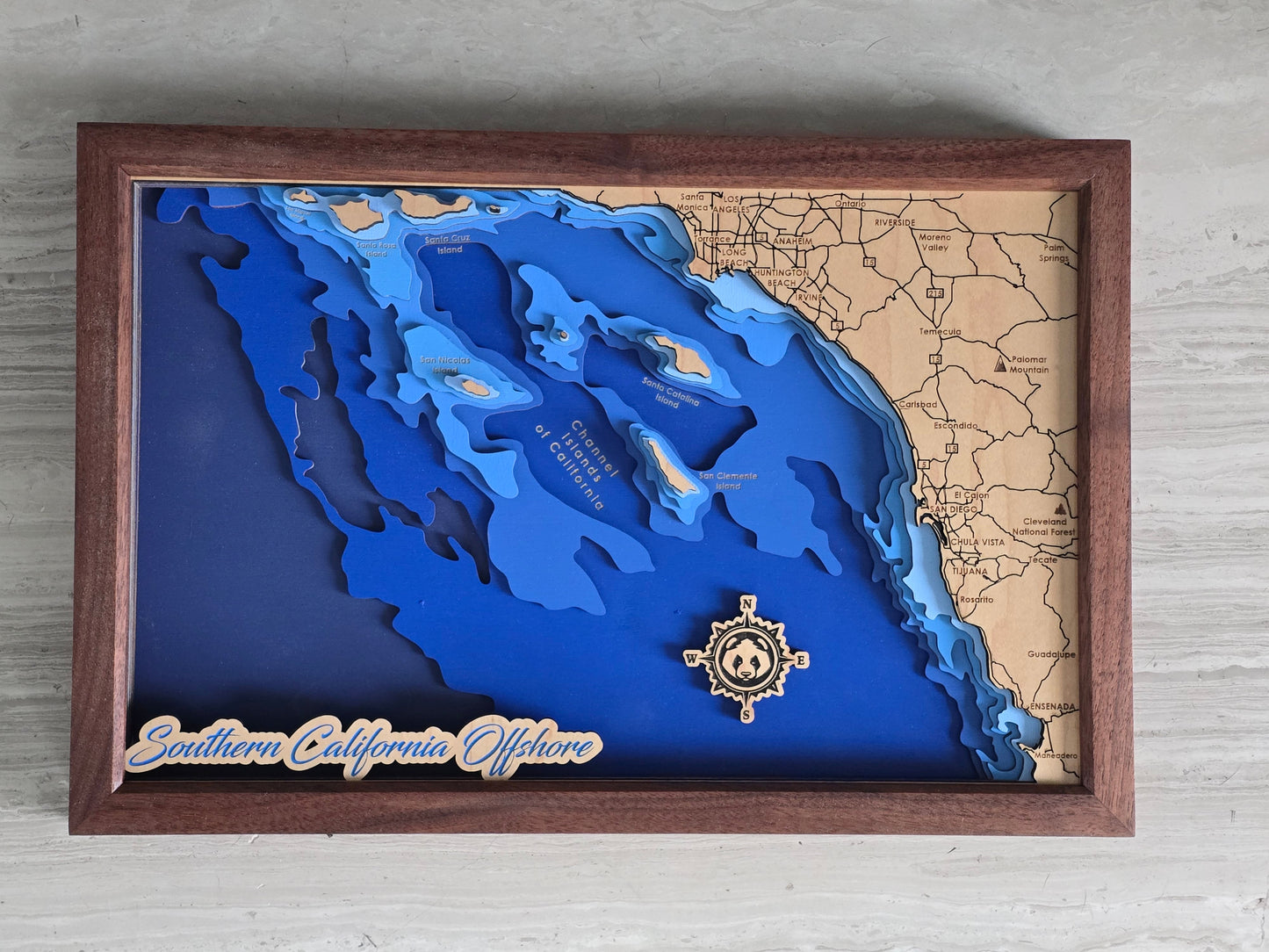 Wooden framed map with blue water and brown landmasses, featuring a decorative emblem on a light gray background.