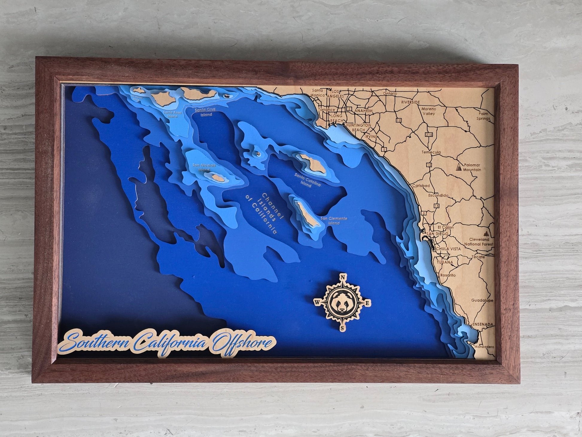 Wooden framed map with blue water and brown landmasses, featuring a decorative emblem on a light gray background.